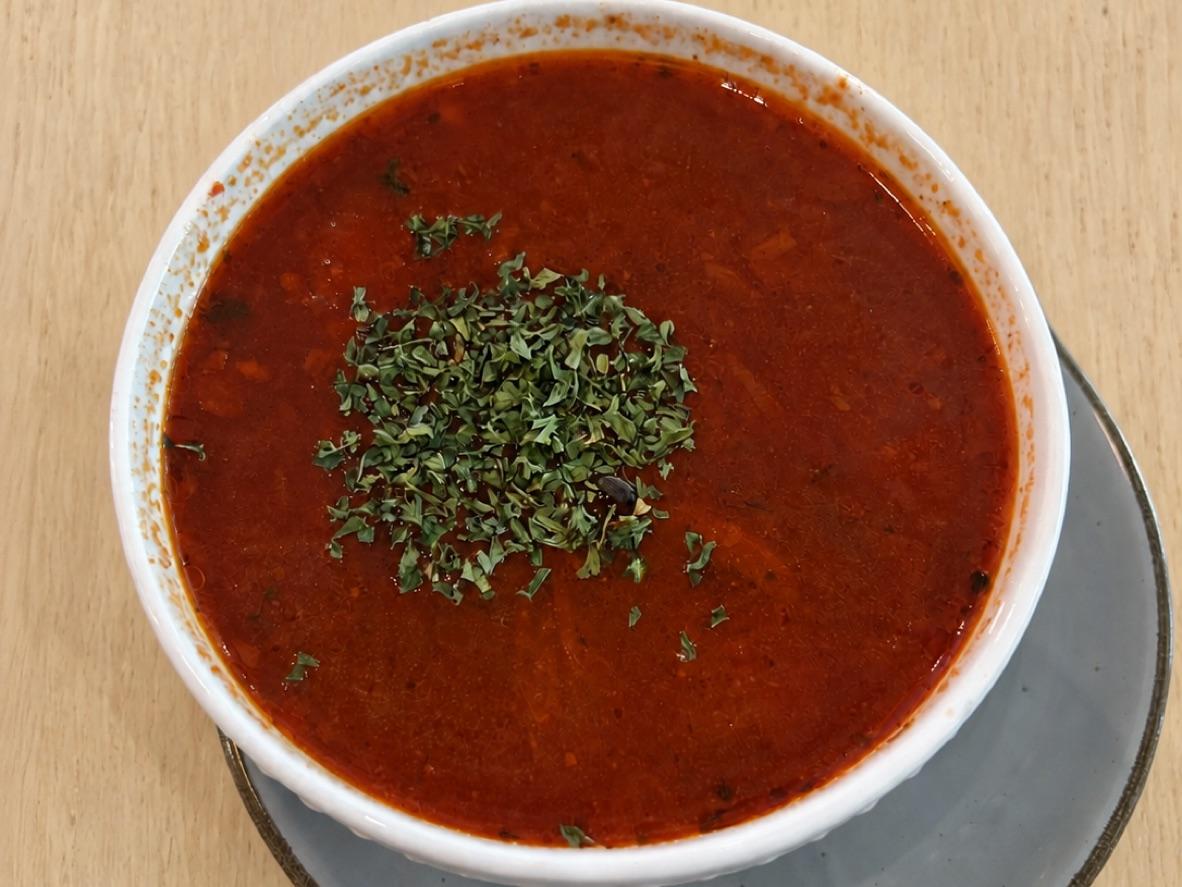 A white bowl of red Ukrainian borscht topped with herbs on a wooden table
